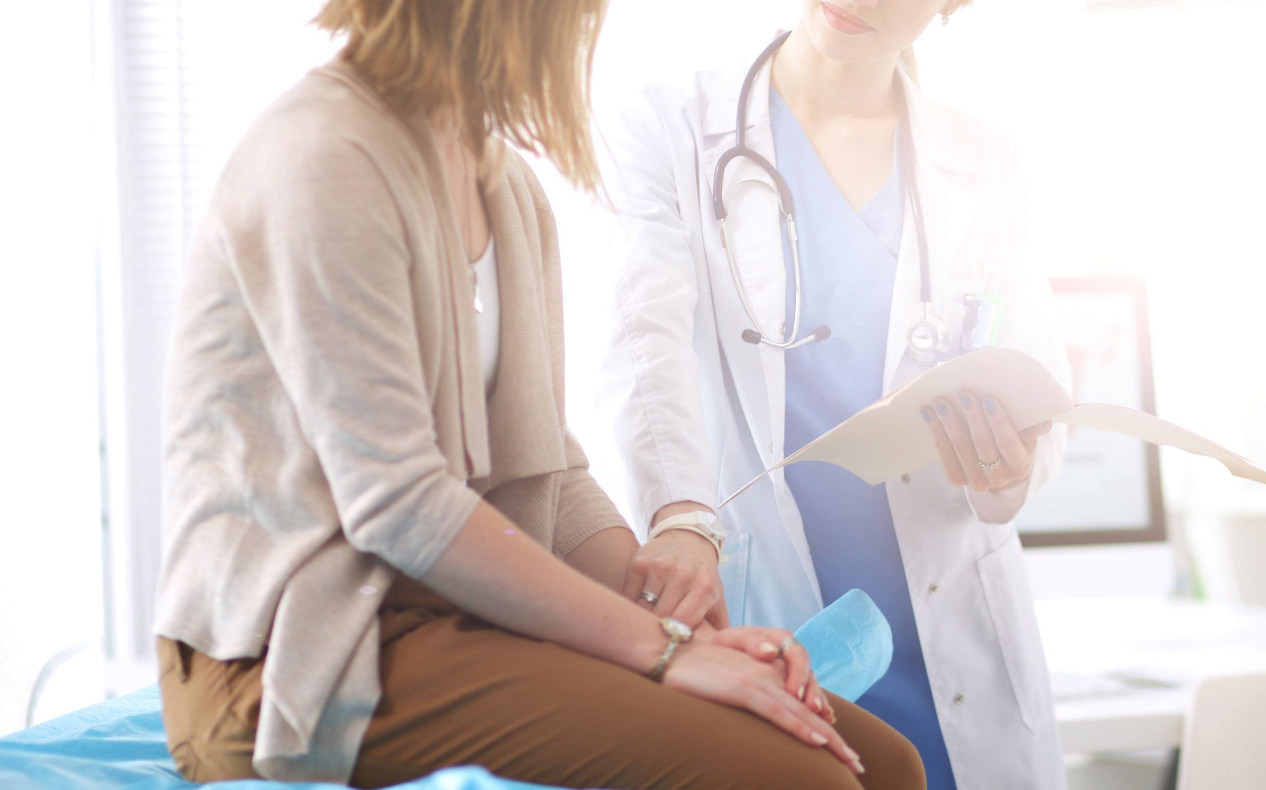 A female doctor in a blue scrub holding a document while discussing public health programs with a female patient sitting on an examination table in a sunlit medical office.