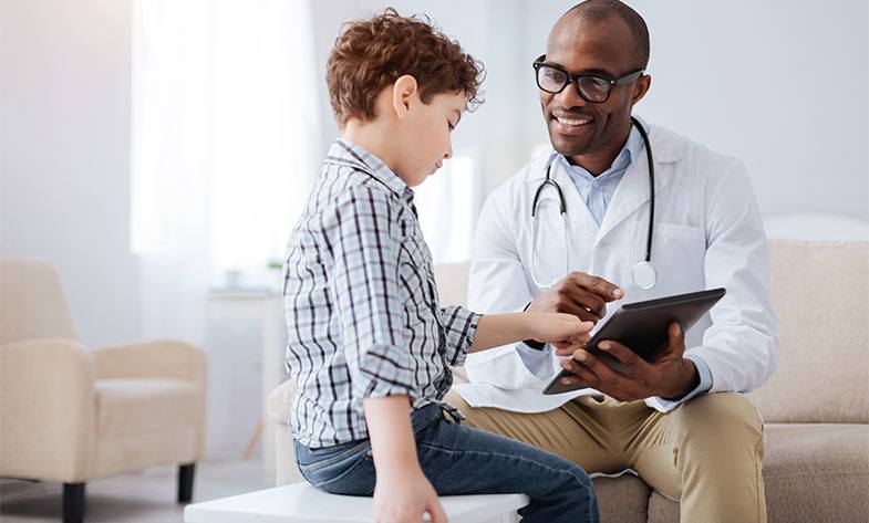 A young boy in a striped shirt smiles while looking at a digital tablet held by a smiling male doctor in glasses and a white coat, sitting in a bright medical office involved in public health programs.