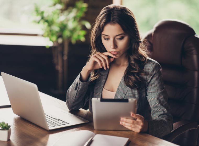 A professional woman in a plaid blazer sits at a desk, intently focusing on a tablet while a laptop is open beside her in a brightly-lit office.