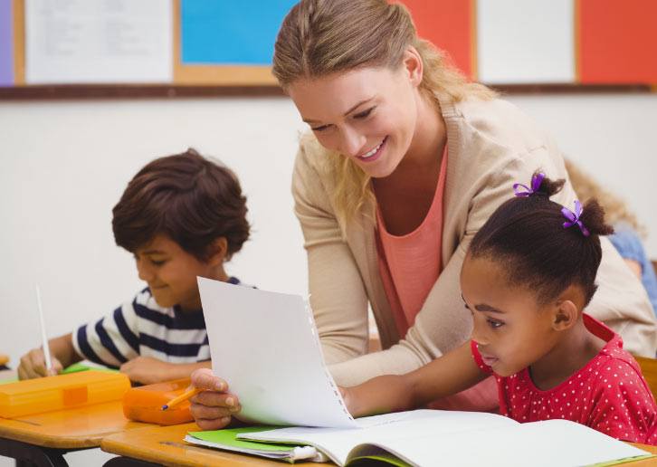 A teacher smiles while helping a young girl with her early childhood education schoolwork in a vibrant classroom setting, as other students work independently in the background.