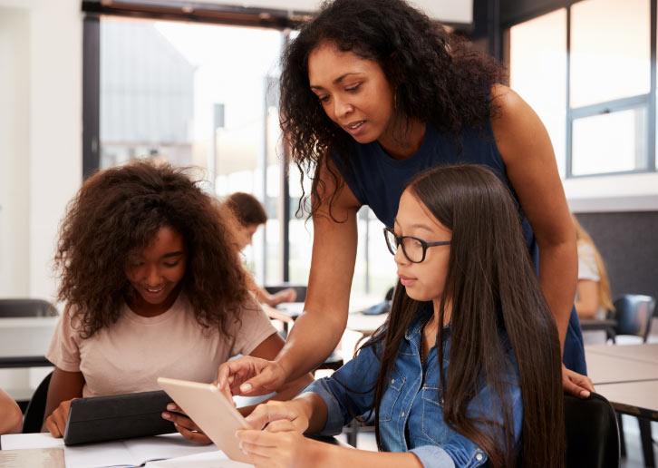 An educator leans over to assist two female students using a tablet in an education-focused classroom, with other students working in the background.