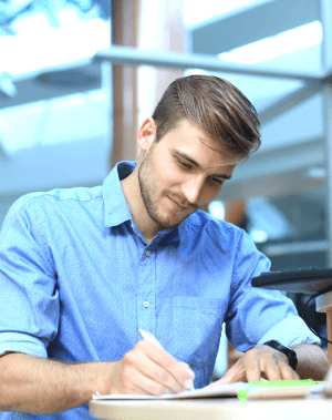 A focused social worker in a blue shirt writing in a notebook at a desk with windows in the background.