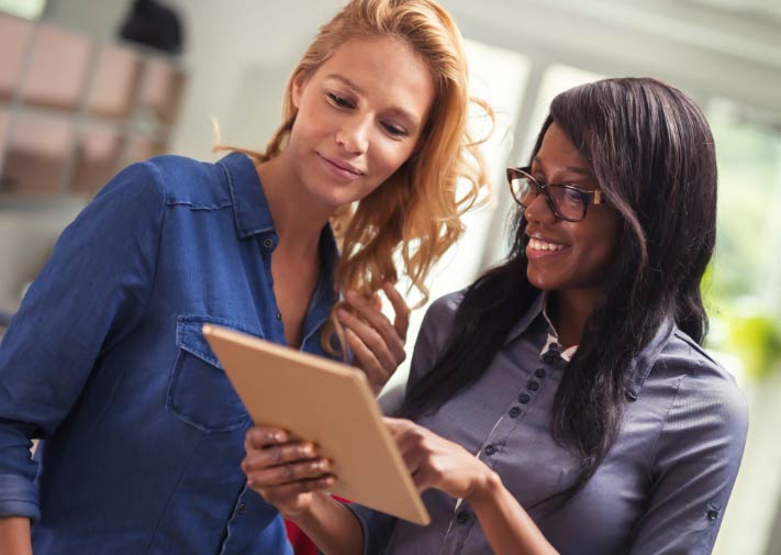 Two social workers, one Black and one Caucasian, smiling and looking at a tablet together in a modern office environment.