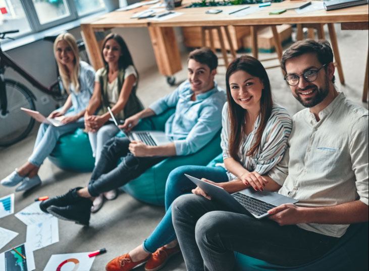 A group of five young professionals, including one with a Doctorate in Psychology (PsyD), sitting on bean bags, smiling at the camera, holding laptops and documents in a casual office environment.