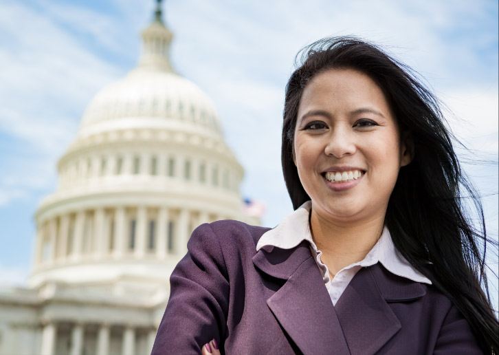 A smiling woman with a Master's in Public Administration stands confidently in professional attire in front of the United States Capitol building under a partly cloudy sky.