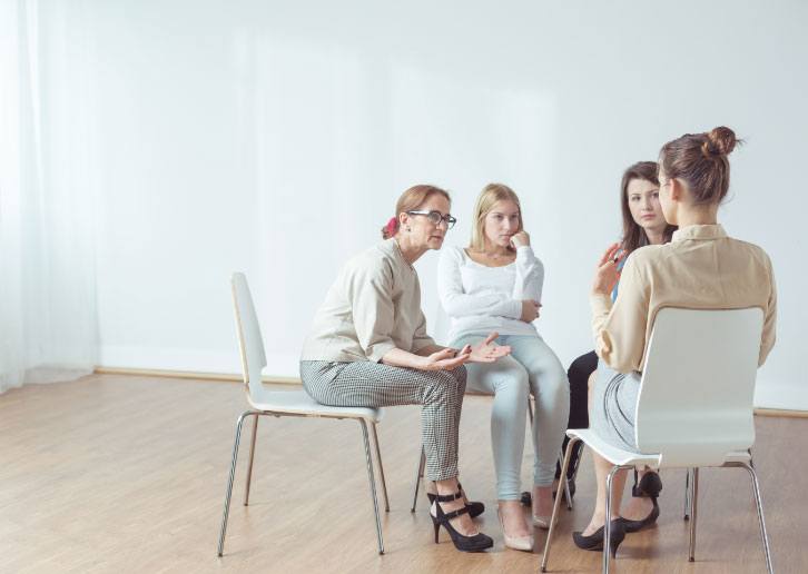 Four women seated in chairs in a circle, engaged in conversation in a bright, minimalist room within the industry sector. They appear focused, with one gesturing while talking.