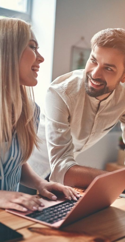 A man and a woman smile at each other while substitute together on a laptop in a warmly-lit room, suggesting a collaborative and friendly environment.