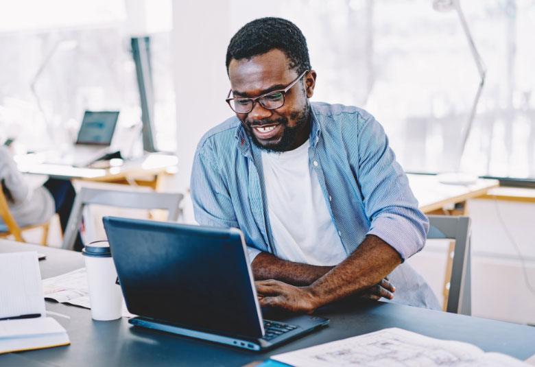 A cheerful man wearing glasses and a light blue shirt, working on a laptop at a busy cafe table strewn with papers and books related to psychology programs.