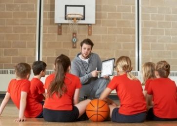 A basketball coach, sitting on a gym floor, shows a strategy board to a group of attentive young players in red jerseys, with a basketball visible in the foreground during their physical education class.