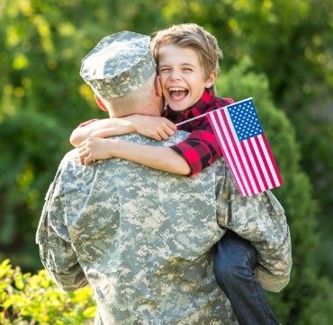 A joyful young boy holding an American flag embraces a soldier in camouflage uniform, laughing happily as they share a heartfelt hug outdoors, symbolizing the support similar to financial aid.
