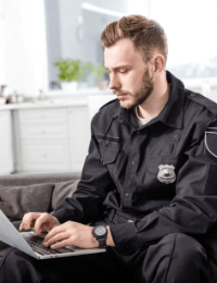 Certainly! Here is the modified description using a keyword: A police officer in uniform, focused on typing on a laptop while sitting indoors with a bright, blurred background of the police station.