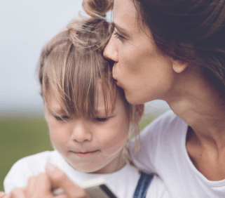 A woman affectionately kissing a young child on the head while the child looks at a smartphone. They are outdoors with a blurry green background, symbolizing a moment of love and care in an anti-bullying awareness context.