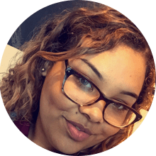 A close-up portrait of a young woman with curly hair wearing glasses and smiling at the camera, symbolic of homeless students overcoming challenges in higher education.