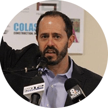 A man with a beard and mustache, wearing a blue shirt and dark blazer, speaks at a podium with microphones from SNLZ and KGW, against a backdrop with affordable housing assistance logos.