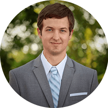 A professional portrait of a man wearing a gray suit, light blue shirt, and striped tie, smiling gently against a blurred green background, featured in an online guide.