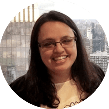 A smiling woman with long dark hair and glasses, wearing a white T-shirt, stands indoors in front of a window overlooking skyscrapers, holding a copy of the "Beginners Guide to Praxis.