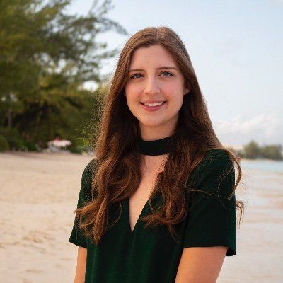A young woman with long brown hair and a green top, pursuing a career in social justice, smiles gently at the camera, standing on a sandy beach with trees and a clear sky in the background.