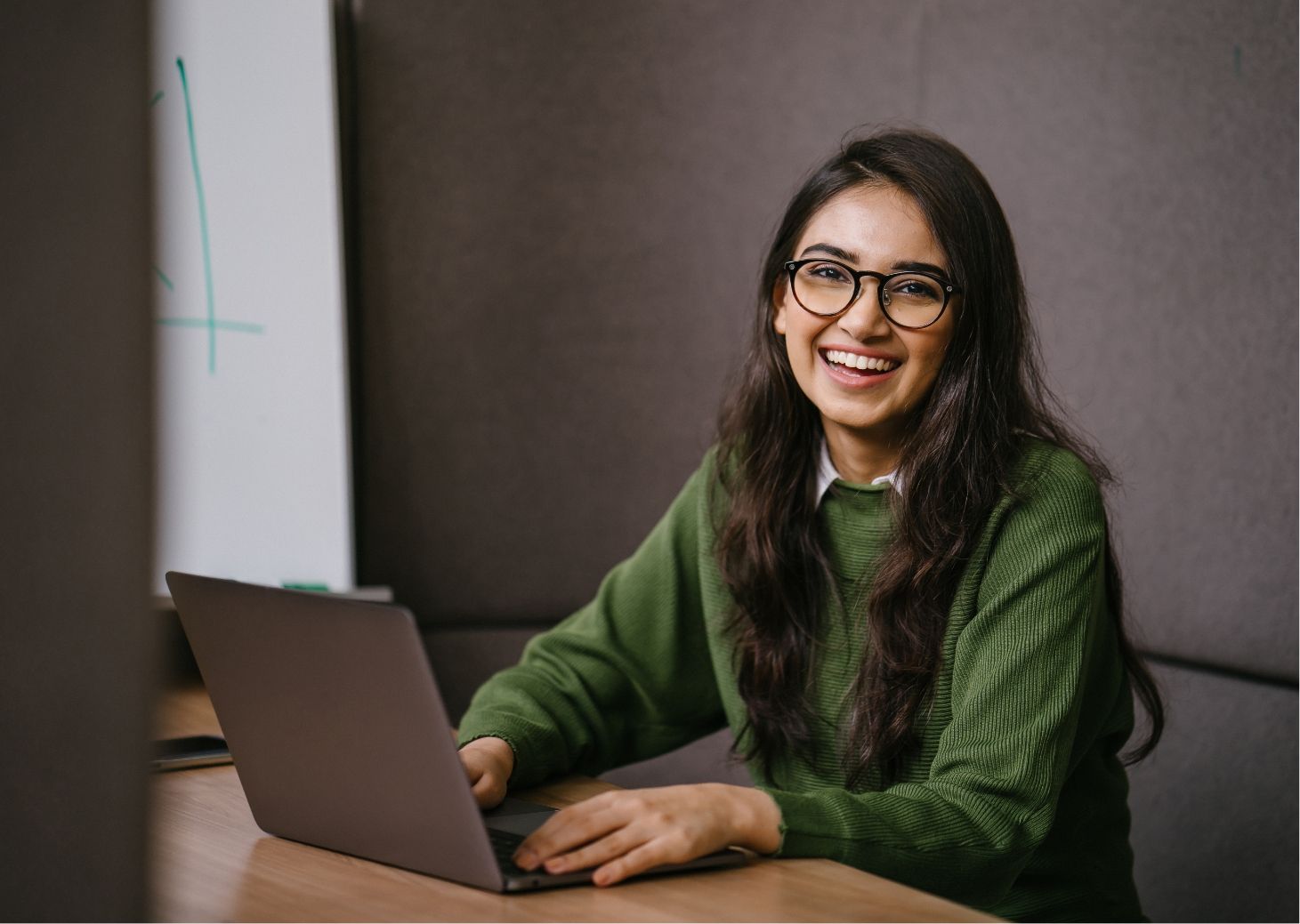 A smiling woman with glasses, wearing a green sweater, sits at a desk working on a laptop, with educational materials on the whiteboard in the background.