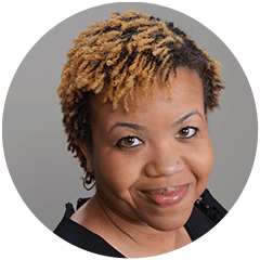 A smiling woman with two-toned curly hair, featuring dark roots and blonde tips, against a neutral background. She wears a black top and has a friendly expression.