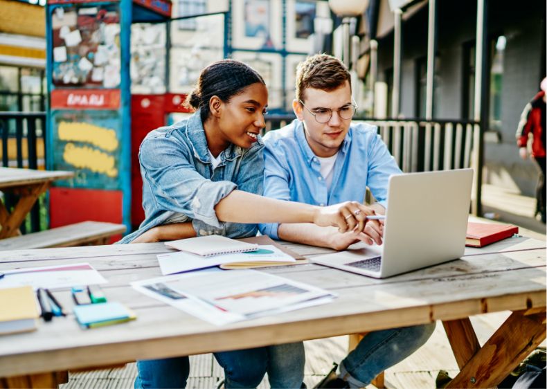 Two young adults, one woman and one man, sit at a wooden table outdoors, working together on a laptop surrounded by notebooks and pens. They are discussing their college courses and pointing at the laptop screen.