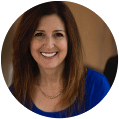 A smiling woman with shoulder-length brown hair wearing a blue blouse and a gold necklace, posing for a portrait.