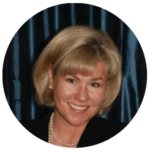 A smiling woman with short blonde hair, wearing a dark blazer and a pearl necklace, against a dark blue curtain background, possibly pondering her college majors.