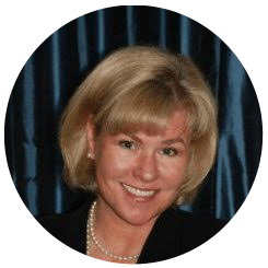 A smiling woman with short blonde hair, wearing a dark blazer and a pearl necklace, against a dark blue curtain background, possibly pondering her college majors.