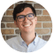 Profile photo of a smiling online student with glasses, wearing a white shirt, standing in front of a brick wall.