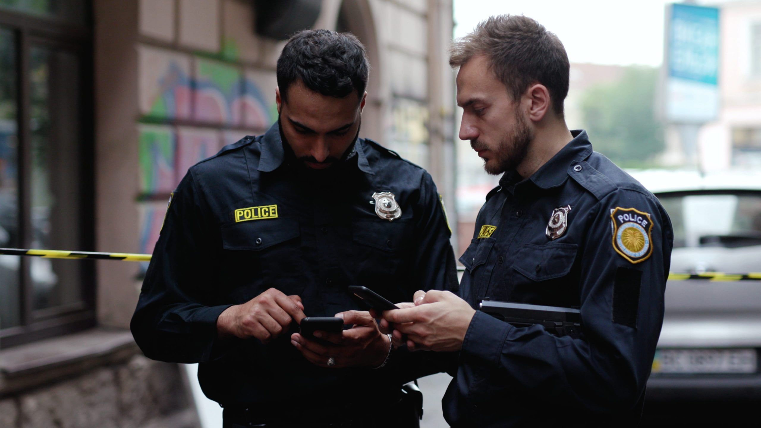 Two law enforcement officers, one holding a smartphone and the other pointing at its screen, standing on a city street behind yellow police tape.