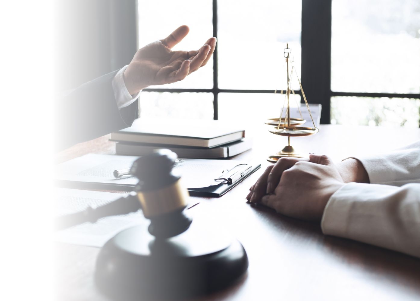 A lawyer's hands are visible on a desk, gesturing during a discussion. A gavel, legal books, and a scale of justice are also on the table, indicating a legal setting.