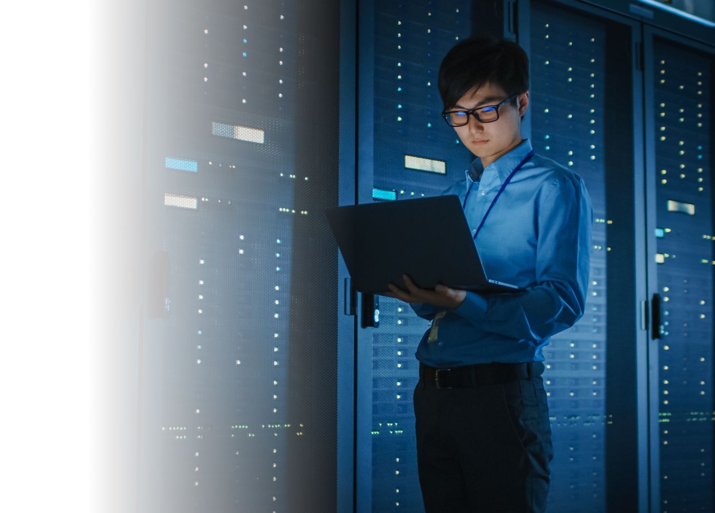 A person in a blue shirt and glasses stands in a server room, intently examining a laptop with illuminated server racks in the background.