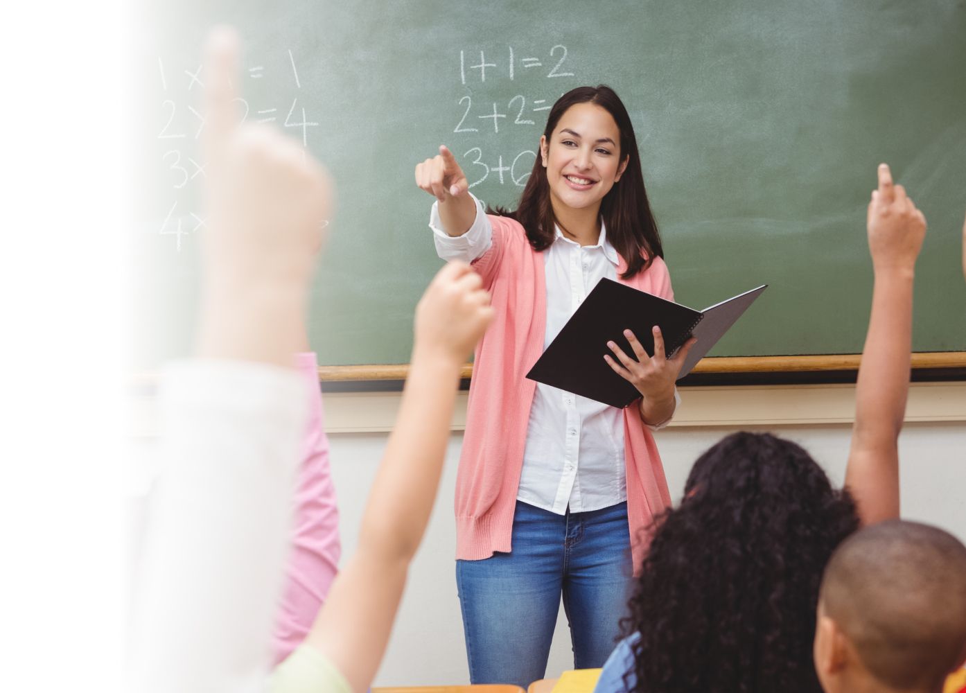 A smiling female teacher holding a book points towards a student in a classroom with students raising their hands, with math problems written on the blackboard behind.