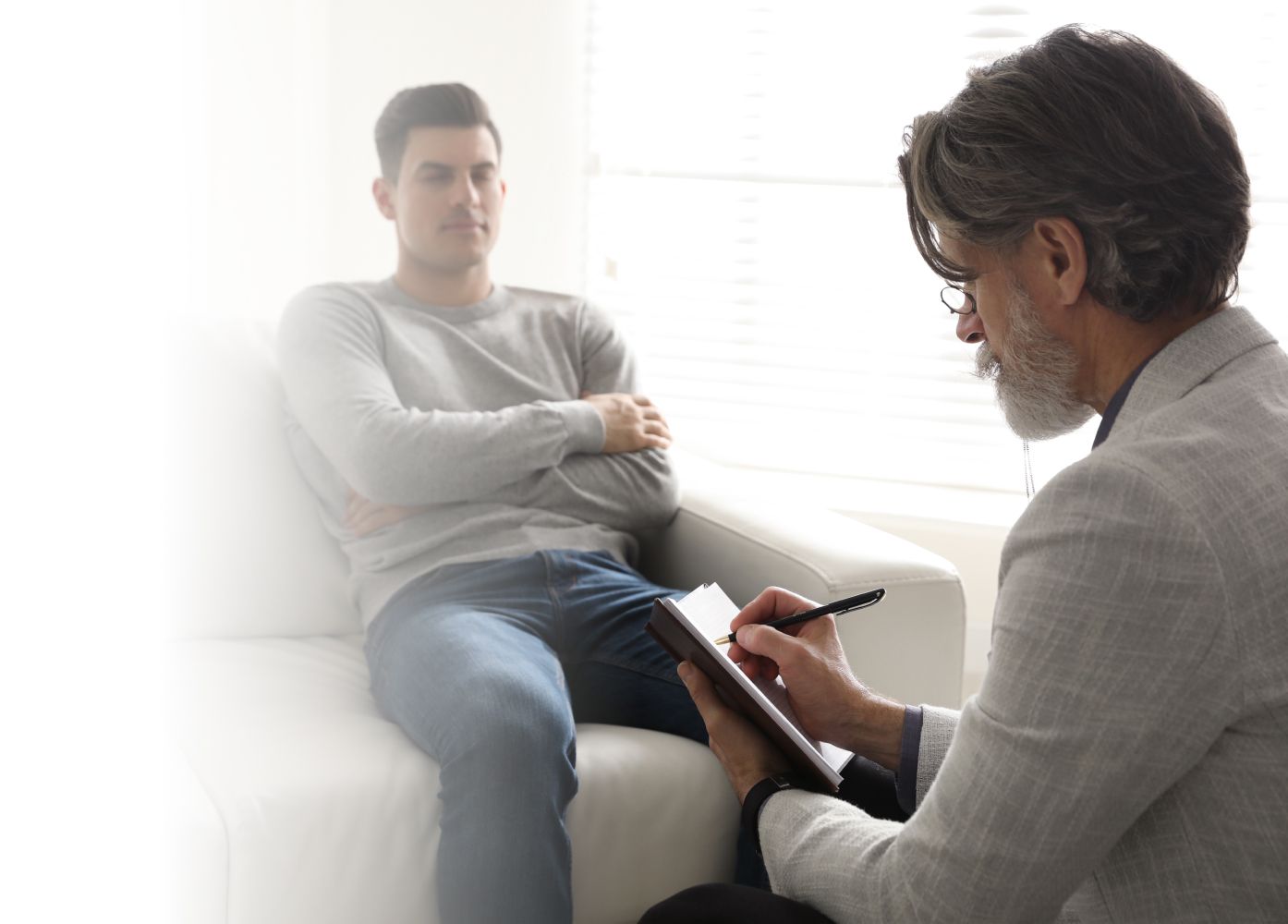 A young man sits with crossed arms on a couch, looking contemplative. An older man sits opposite him, taking notes on a clipboard. They are in a bright, sunlit room.