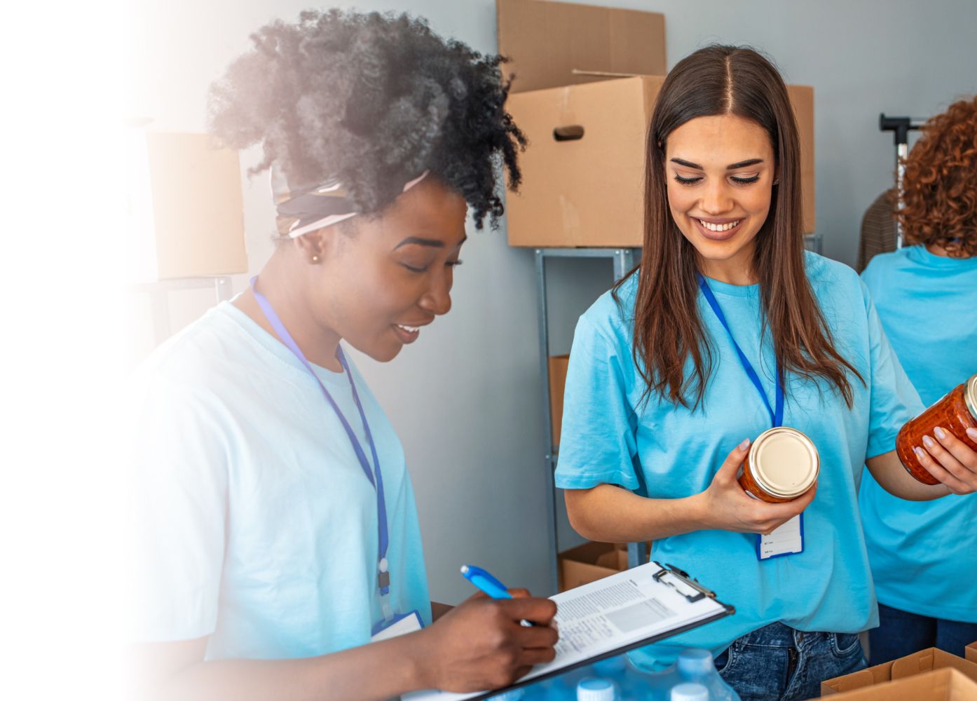 Two women volunteers in blue t-shirts working at a food drive, with one recording details on a clipboard while the other holds a food jar.