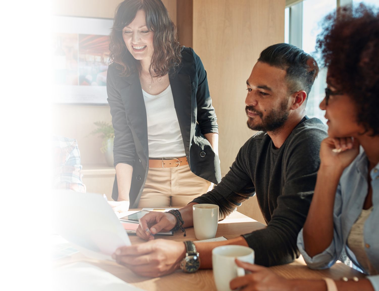 Three professionals in a sunlit meeting room discuss over a laptop, showing engagement and teamwork, with one woman standing and two seated colleagues.