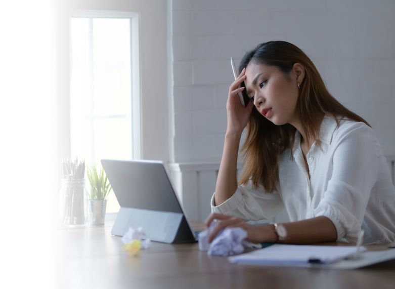 A woman appears thoughtful while working on a laptop at a desk with crumpled paper nearby, signaling recent failure, and gazing at the screen with her head resting on her hand.