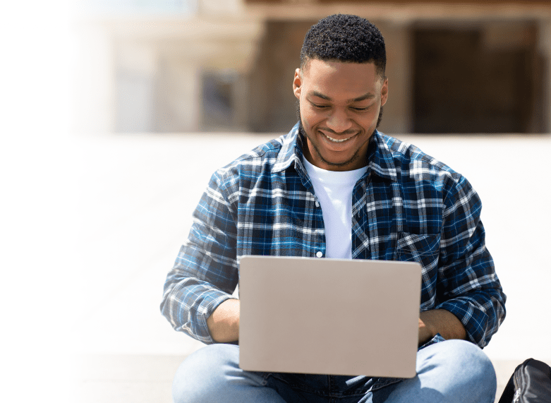 A cheerful young man sitting on steps outdoors, engaging in online education using a laptop. He is dressed in a blue and white plaid shirt over a white T-shirt.