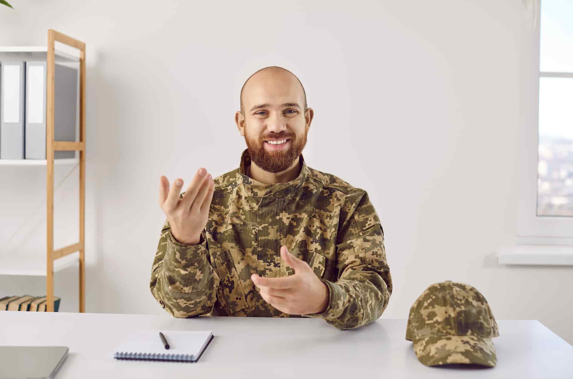 A smiling man in military camouflage uniform sitting at a desk, gesturing with his hands as if explaining something, with a notebook on the side and a cap on the table.