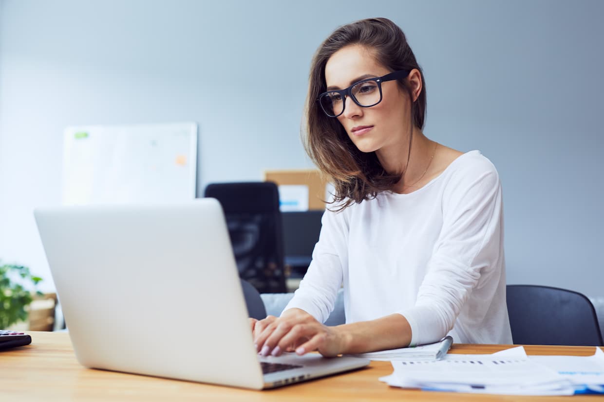 A professional woman wearing glasses and a white top works intently on evaluating student loan risk at a desk, surrounded by papers, in a well-lit office space.