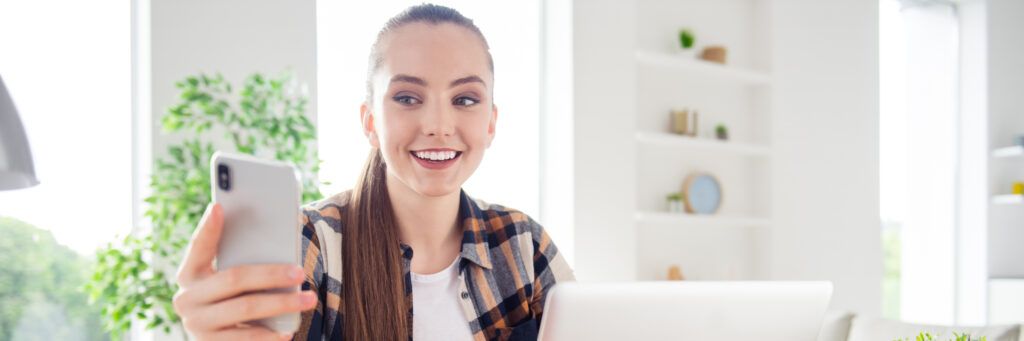 A female student sitting at a desk with a laptop in front of her while holding a cell phone.
