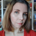 A woman with shoulder-length brown hair and fair skin, wearing a red top, stands in front of a bookshelf filled with various books. She looks directly at the camera with a subtle expression.