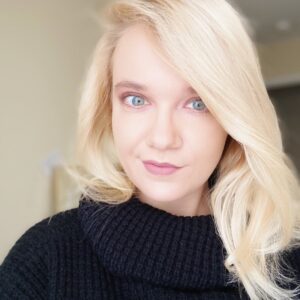 A close-up portrait of a woman with blonde hair, blue eyes, and light skin, wearing a black turtleneck sweater and subtly smiling at the camera.