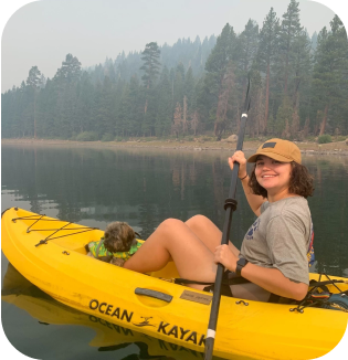 A woman in a yellow kayak smiles, holding a paddle on a serene, misty lake. She wears a cap, and a dog peeks from the kayak. Trees in soft focus stand in the background.