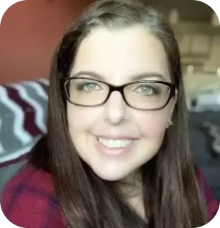 A close-up portrait of a smiling woman with long brown hair and glasses, wearing a red blouse, indoors with soft-focus background.