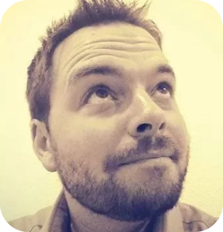 A sepia-toned close-up photo of a smiling man with short hair and a trimmed beard, looking upwards to his right, against a plain background.