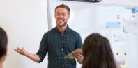 A smiling male teacher holding a tablet while engaging with students in a bright classroom setting, standing in front of a whiteboard.