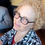 A woman with curly blonde hair and glasses smiling slightly at the camera, wearing a black and white patterned top. She is in a well-lit indoor setting.