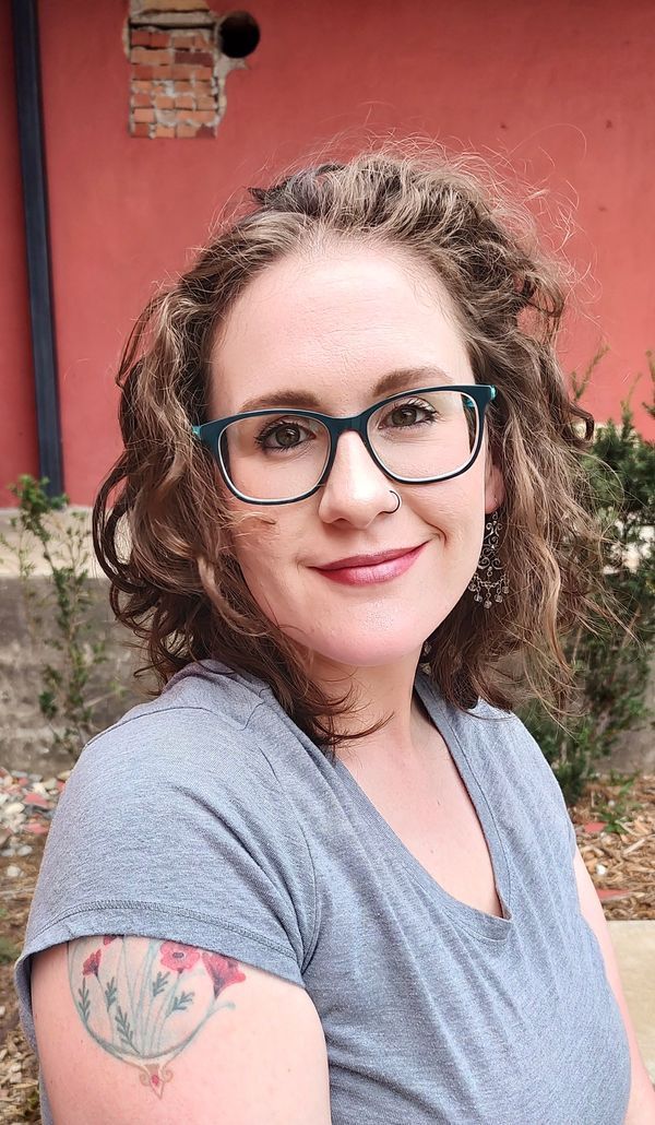 A woman with curly hair and glasses smiles at the camera, standing against a red wall. She has a floral tattoo on her arm.
