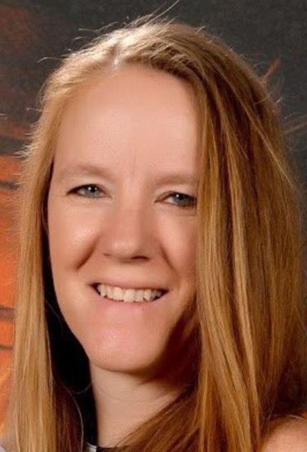 A close-up portrait of a smiling woman with long, straight blonde hair and fair skin, set against a blurred brown background.