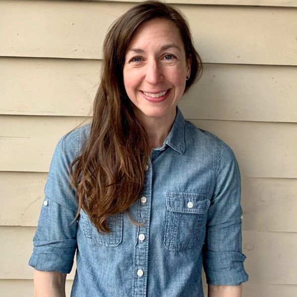 A smiling woman with long brown hair wearing a denim shirt stands in front of a beige siding background.